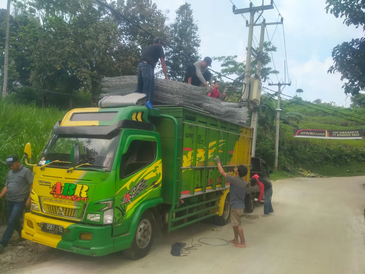 Truk hijau mengangkut bronjong kawat melewati jalan desa